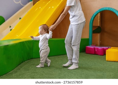Baby taking first steps while being supported by an older child in a daycare play area, showing care, balance training, and early development.