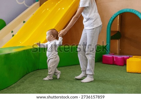 Baby taking first steps while being supported by an older child in a daycare play area, showing care, balance training, and early development.