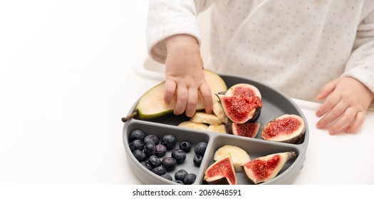 Baby Taking Fruits And Berries From Silicone Plate On White Background, Close-up Shot. Child Self Nutrition Concept. Toddler Eating Fig,pear,blueberries With Hands.Copy Space.Selective Focus