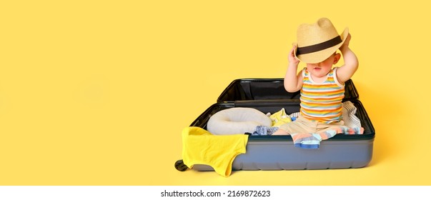 Baby Toddler Boy Sits Inside A Suitcase With Clothes, Studio Yellow Background. A Child Plays In A Big Bag While Packing Luggage For A Vacation Trip, Copy Space. Kid Age One Year (12 Months)