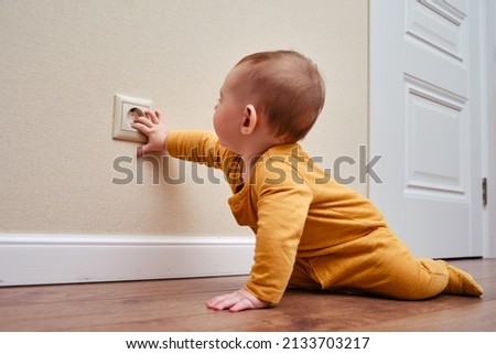 Baby toddler reaches into the electrical outlet on the home wall with his hand. Danger and protection of child fingers from electric shock, aged 6-11 months