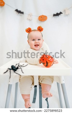 The baby toddler is sitting behind a high chair surrounded by Halloween attributes: pumpkins, spiders and garlands. Halloween Concept