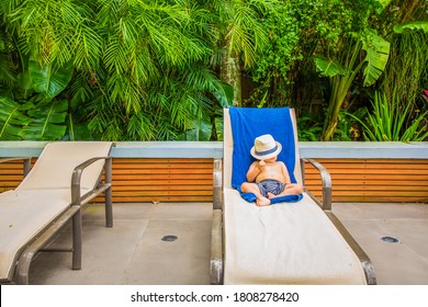 Baby Toddler Small Child Laying Out On A Lounger Chair Lounger Outdoors Poolside Tanning Under The Sun Wearing Swim Trunks Swimsuit And A Fedora Hat Which He Brings Down To Cover His Face With