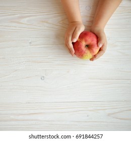 Baby Two Hands Hold Apple On White Wooden Background. Toddler's Hands And Fresh Fruit Top View. Flat Lay Diet And Healthy Food Concept.