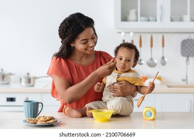 Baby Weaning. Caring Black Mother Feeding Little Toddler Son From Spoon In Kitchen, Loving African American Mommy Giving Porridge Or Mash Fruit Puree To Cute Little Child At Home, Closeup Shot