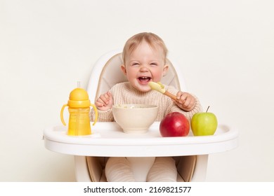 Baby wearing knitted sweater sitting in high chair and feels hungry, holding spoon and eating puree or porridge, enjoying apples and water in yellow bottle, posing isolated on white background. - Shutterstock ID 2169821577