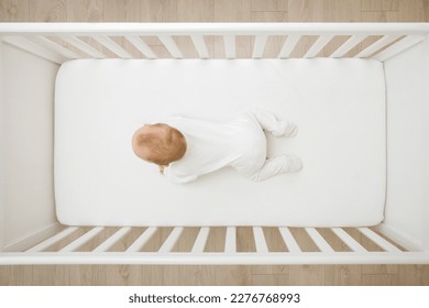 Baby in white bodysuit try crawling on knee and arms on mattress in wooden crib at home room. Top view. 5 to 6 months old infant development.