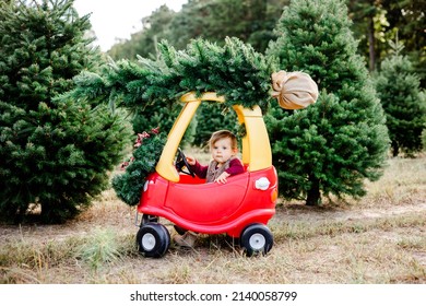 Baby Young Girl Toddler Picking Out Christmas Tree At Christmas Tree Farm Driving Toy Kid Car With Christmas Tree On Top Of Car In Winter For The Holiday