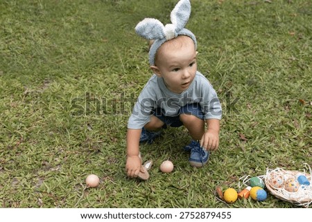 Baby's First Easter Egg Hunt in Bunny Ears. An adorable toddler in bunny ears is joyfully searching for colorful Easter eggs scattered on the green grass. This charming image captures the excitement of a child participating in a festive Easter egg hunt outdoors.