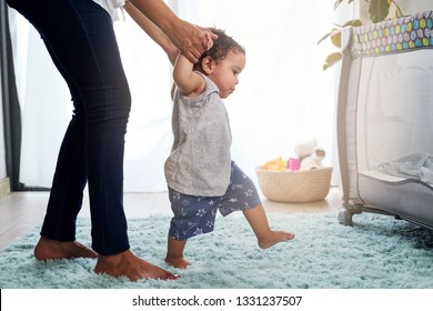 Die ersten Schritte des Babys, die die Hände der Mutter halten, süßes instabiles Laufen in der Heimkinderei mit Kinderbett