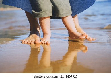 Baby's First Steps. Mother And Small Child Walking Barefoot On Beach Sand Near By Ocean. Happy Young Family In Nature. Mother And Baby Playing Outdoors On Sea Beach. Close-up Human Feet.