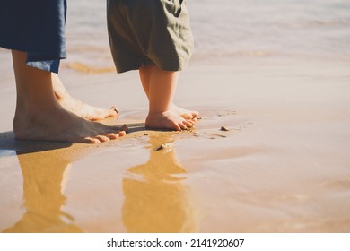 Baby's First Steps. Mother And Small Child Walking Barefoot On Beach Sand Near By Ocean. Happy Young Family In Nature. Mother And Baby Playing Outdoors On Sea Beach. Close-up Human Feet.