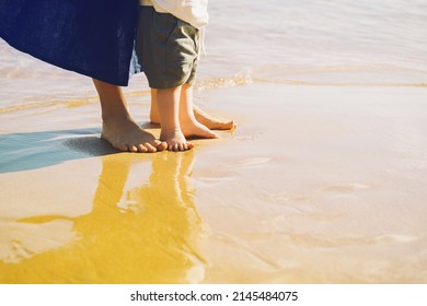 Baby's First Steps. Mother And Small Child Walking Barefoot On Beach Sand Near By Ocean. Happy Young Family In Nature. Mother And Baby Playing Outdoors On Sea Beach. Close-up Human Feet.