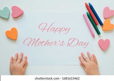 Baby's Hands On White Sheet Of Paper. Happy Mother's Day Greeting Card. Drawing With Pencil Crayons. Children Art Concept. Handmade Mother’s Day Craft. Top View, Flat Lay. 