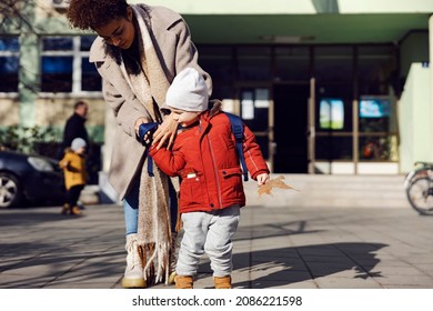 Babysitter Putting Backpack On Kindergarten Child In The Schoolyard. Babysitter Standing In The Schoolyard With Kindergarten Child And Putting A Backpack On His Back.