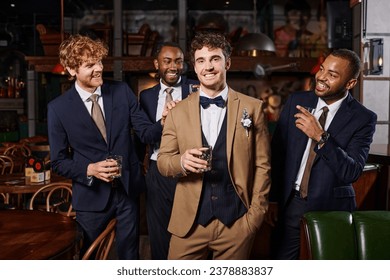 bachelor party, happy interracial best men looking at groom in suit standing with whiskey in bar