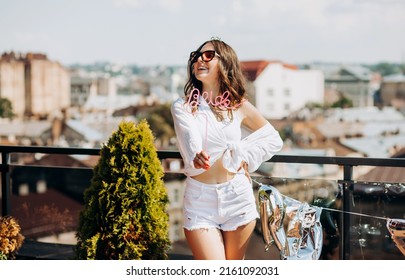 Bachelorette Party. Happy Future Bride In A White Shirt And Shorts Poses On The Roof Of The House Overlooking The City.