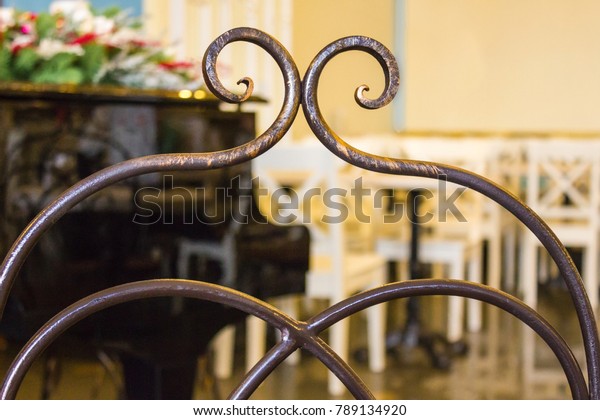 Back of an ancient chair made of forged bronze,\
handicraft production. In the background there is a grand piano and\
white wooden furniture. Vintage interior of the cafe. Morning,\
natural light.