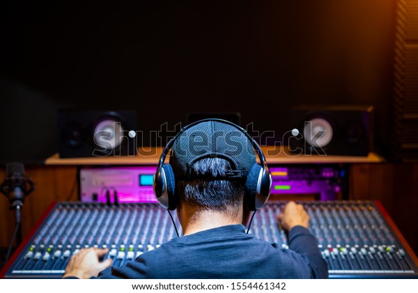 back of asian male professional\
music producer, sound engineer mixing a song on audio mixing\
console in recording studio. music production, post production\
concept