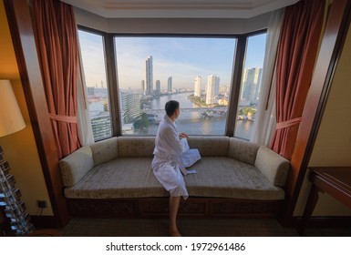 Back of Asian man sitting on sofa couch with window glass in hotel room with Bangkok City skyline in Thailand. Skyscraper buildings. Downtown skyline. Urban town. Relaxing area. People lifestyle.