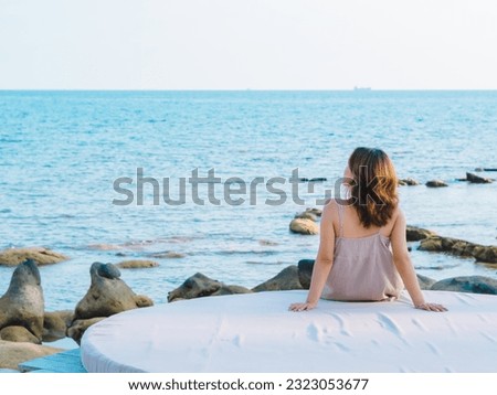 Back of Asian woman in casual dress sitting armrest on cushion in front of the Seascape beach in summer vibes. Lonely female relaxing smile at the sea and looking far away. Holiday travel vacation.