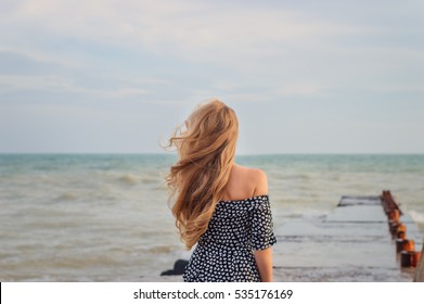 back beautiful slim girl with long flowing blond hair in the wind against the background of the sea and horizon. concept expectations, light sadness


