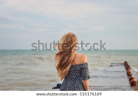 back beautiful slim girl with long flowing blond hair in the wind against the background of the sea and horizon. concept expectations, light sadness


