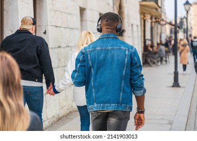 The back of a black man in a denim jacket, jeans with black wireless headphones among passers-by on the street on a sunny day