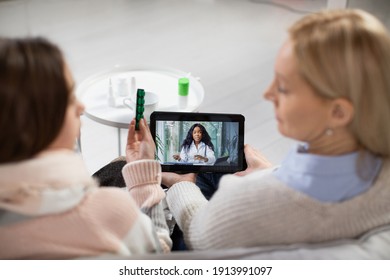 Back Blurred View Of Caring Mother And Sick Teen Daughter, Sitting With Tablet Pc And Listening Instructions From Their Family African Female Doctor About Treatment Via Video Call. Focus On Tablet