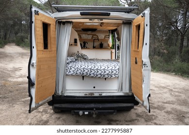 Back of camper with open doors, interior with wooden bed and kitchen, wooden doors, heart quilt and grey curtains, large white camper van, lonely forest interior.