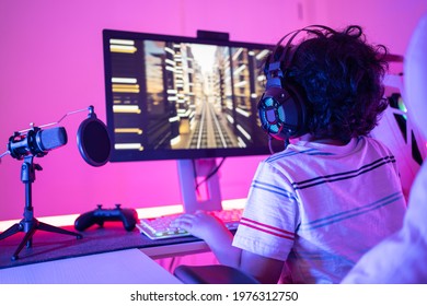 Back Of Cute Curly-haired Little Asian Boy Gamer Wearing Headphones Sitting Playing Video Games On A Personal Computer With RGB Keyboard Next To Joystick And Desktop Microphone In A Violet Neon Room.