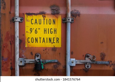 The Back Door Of An Aged And Rusty Heavy-duty Red Container With Locks And Sealed Padlock. Yellow Warning Sign About High Container Height Is Marked On The Door