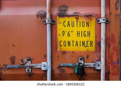 The back door of an aged and rusty heavy-duty red container with locks and sealed padlock. Yellow warning sign about high container height is marked on the door