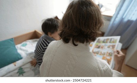 Back of elderly woman reading a book to her young grandson, seated on a bed. The room is bright with natural light, boy listening attentively to grandmother's storytelling - Powered by Shutterstock