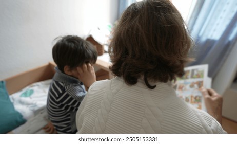 Back of elderly woman reading a book to her young grandson, seated on a bed. The room is bright with natural light, boy listening attentively to grandmother's storytelling - Powered by Shutterstock