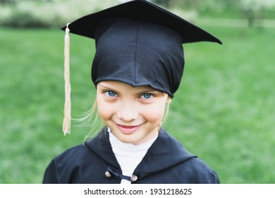 Back to elementary school concept. Little girl goes to first grade. Ceremony of graduating. Black gown, academic cap with tassels. Party with parents, teachers in park, yard. Last day, end of year.