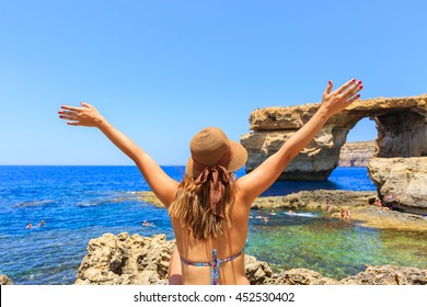 Back of female tourist wears hat, bikini, raises hands up and enjoys summertime with background of Azure window, famous tourist attraction on Gozo island, Mediterranean sea, Malta