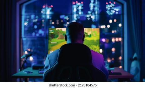 Back Of The Head Angle Of A Successful African American Gamer Sitting In Front Of Big Window And Playing A Video Game On Personal Computer In A Neon Lit Living Room At Home. Young Man In Victorious.