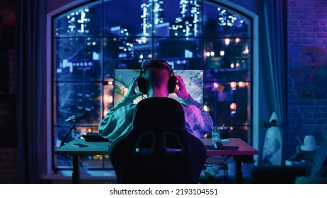 Back Of The Head Angle Of A Successful Stylish Gamer Putting On Headphones And Playing A Video Game On Personal Computer In A Neon Lit Living Room At Home. Young Man In Victorious.