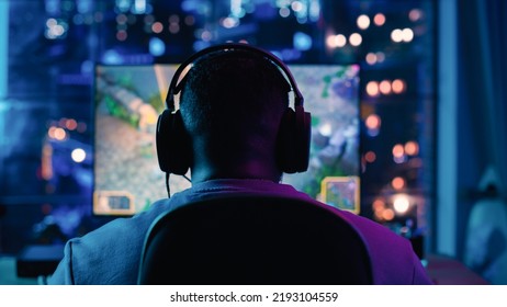 Back Of The Head Shot Of An African American Gamer Wearing Headphones And Playing A Video Game On Personal Computer In A Neon Lit Living Room At Home. Cozy Evening At Home In Loft Apartment.