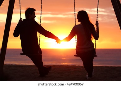 Back light portrait of a couple silhouette sitting on swing holding hands watching a sunrise on the beach with the sun in a warmth background