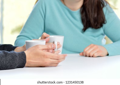 Back light profile of hands of two friends or couple talking holding coffee cups sitting in a table at home with a window in the background