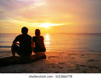 Back of loving couple sitting side by side on tropical beach with dramatic sunset or sunrise sky background. Happy young girlfriend and boyfriend in love on vacation. Valentines Day, loving concept.