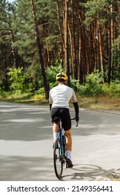 Back Of A Male Cyclist Rides Outside The City On A Bicycle In The Woods On An Asphalt Road On A Background Of Trees. Vertical