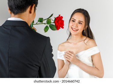 Back Of Man Wearing Black Tuxedo Giving Rose To Young Attractive Asian Woman, Soon To Be Bride And Groom, Woman Wearing White Wedding Gown. Concept For Pre Wedding Photography.