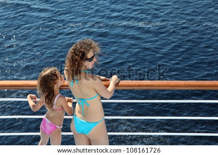 Back of mother and daughter dressed in swimsuits standing on deck of ship and looking into distance