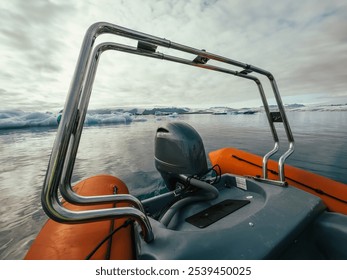 The back of an orange motorized boat floats on calm, icy waters surrounded by drifting icebergs under a cloudy sky. The metal frame and engine add a modern contrast to the serene, frozen landscape. - Powered by Shutterstock