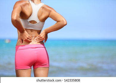 Rückenschmerzen. Athletische Frau in rosafarbener Sportbekleidung, die am Meer steht und die Muskeln ihres unteren Rückens rutscht, zerschnittenes Torso-Portrait.