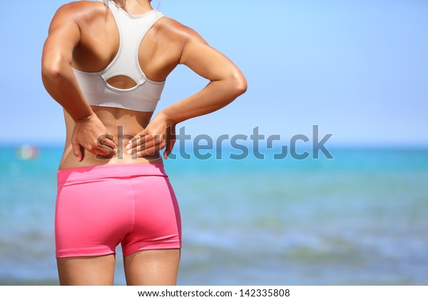 Back pain. Athletic woman in pink sportswear\
standing at the seaside rubbing the muscles of her lower back,\
cropped torso portrait.