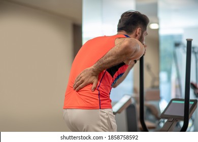 Back Pain. Young Man In Bright Tshirt Exercising On A Gym Machine And Feeling Pain In Back